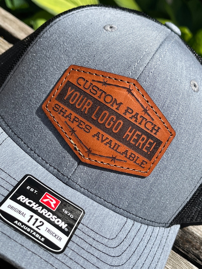 May include: Gray trucker hat with a black mesh back and a brown leather patch. The patch reads "CUSTOM PATCH YOUR LOGO HERE! SHAPES AVAILABLE." The hat also has a Richardson 112 Trucker tag.