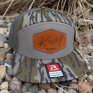 May include: A camouflage patterned trucker hat with a brown leather patch that says "K+M 08.23.2025". The hat has a black mesh back and a red logo that says "Richardson. Flat Bill Snapback."