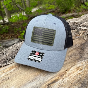 May include: A gray and black trucker hat with a green and black American flag patch on the front. The hat has a black mesh back and a label that reads "Richardson 115 Low Pro Trucker".