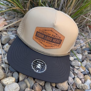 May include: A tan and black baseball cap with a custom patch. The patch is brown with the text "CUSTOM PATCH YOUR LOGO HERE! SHAPES AVAILABLE". The cap has a black brim and a rope detail. A sticker with a helicopter graphic is on the brim.