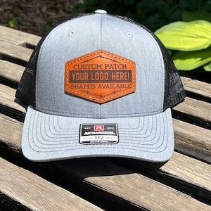 May include: A gray trucker hat with a black mesh back. The hat features a brown leather patch with the text "CUSTOM PATCH YOUR LOGO HERE! SHAPES AVAILABLE". The hat is adjustable and has a Richardson 112 tag.