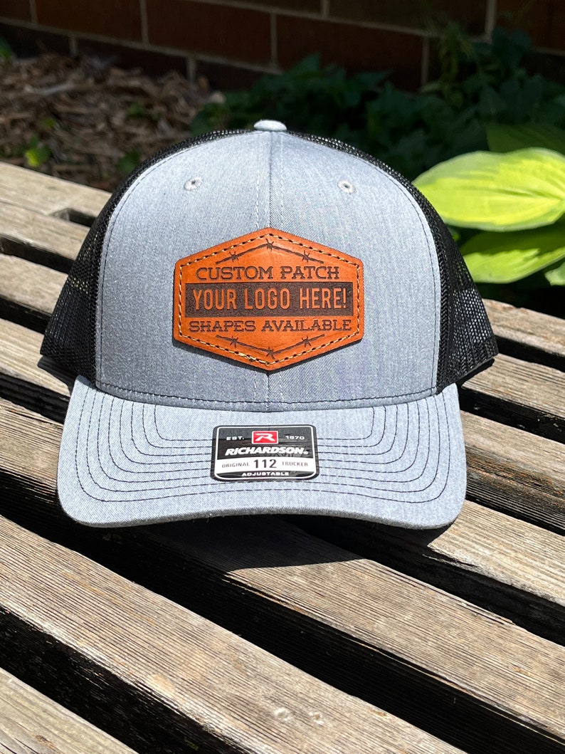 May include: A gray trucker hat with a black mesh back. The hat features a brown leather patch with the text "CUSTOM PATCH YOUR LOGO HERE! SHAPES AVAILABLE". The hat is adjustable and has a Richardson 112 tag.