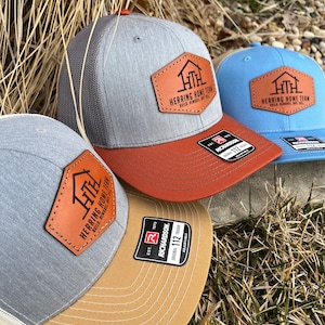 May include: A collection of Richardson 112 trucker hats. The hats feature a gray, brown, and light blue color scheme with a leather patch that reads "Herring Home Team". The hats have a curved brim and a mesh back.