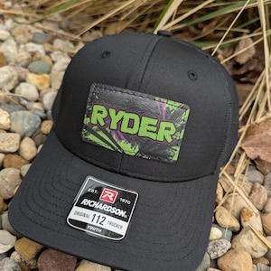 May include: Black youth trucker hat with a green, purple, and black camouflage patch that says "RYDER" in white letters. The hat has a black mesh back and a red, white, and black label that says "EST. 1970 RICHARDSON 112 TRUCKER YOUTH".