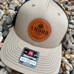 May include: A tan and brown trucker hat with a curved brim. The hat features a leather patch with the text "AMDOR FARMS" and a tractor graphic. A black and white tag reads "RICHARDSON ORIGINAL 112 TRUCKER".