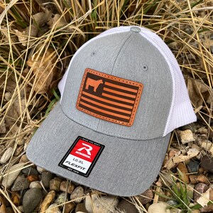 May include: Gray and white trucker hat with a brown leather patch featuring an American flag and a silhouette of a cow. The patch has the text "Wear The Best" on it. The hat has a Richardson logo and the text "LG-XL FlexFit" on the label.