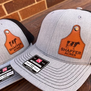 May include: A gray and black trucker hat with a brown leather patch featuring a cow and the text "Shaffer Acres". The hat has a black mesh back and a Richardson 112 label.