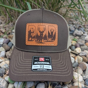 May include: Brown and tan trucker hat with a leather patch that says "DAD" with deer silhouettes. The hat has a curved brim and an adjustable strap. The Richardson brand label is visible.