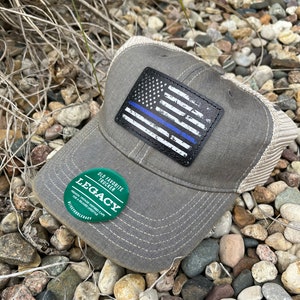 May include: A gray and white trucker hat with a thin blue line American flag patch and a green sticker that says "Old Favorite Trucker Legacy" on the front.