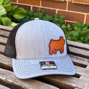 May include: Gray and black trucker hat with a brown leather patch of a pig. The hat has a curved brim and a mesh back. The Richardson brand label is visible on the front.