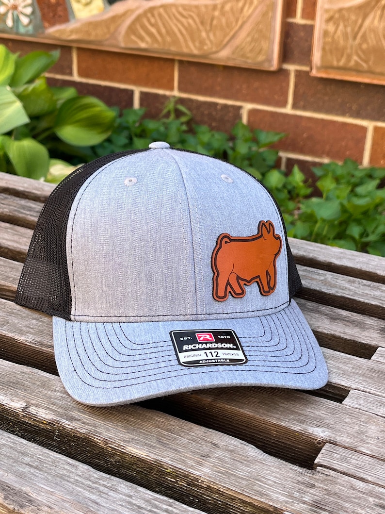 May include: Gray and black trucker hat with a brown leather patch of a pig. The hat has a curved brim and a mesh back. The Richardson brand label is visible on the front.
