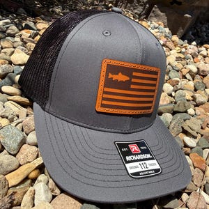 May include: A gray trucker hat with a black mesh back and a rectangular brown leather patch featuring a fish silhouette and horizontal stripes. A black and white sticker on the brim reads: EST. 1970 RICHARDSON ORIGINAL 112 TRUCKER ADJUSTABLE.