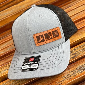 May include: Gray and black youth trucker hat with a brown leather patch featuring a pheasant, antlers, and a fly. The patch has the text "EST. 1970 RICHARDSON. ORIGINAL 112 TRUCKER YOUTH" on the hat.