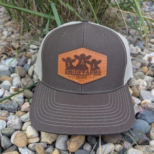 May include: A brown and tan trucker hat with a leather patch. The patch features three cow silhouettes and the text "SMITH FARMS EST.1970". The hat has a curved brim and a mesh back.