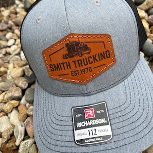May include: A gray and black trucker hat with a brown leather patch that says "Smith Trucking Est. 1970". The hat also has a red and black label that says "Richardson Original 112 Trucker Adjustable".