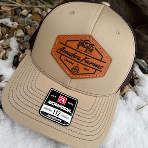 May include: A tan and brown trucker hat with a leather patch featuring the words "Ander Farms" and a cow graphic. The hat has a curved brim and a mesh back. The Richardson tag reads "Original 112 Trucker".