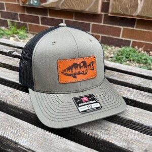 May include: A Richardson 112 trucker hat with a gray crown and brim, black mesh back, and a leather patch featuring a fish and mountain scene. The hat is adjustable.