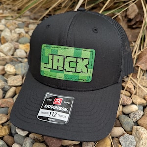 May include: Black trucker hat with a green and white pixelated patch that says "JACK" on it. The hat has a black mesh back and a label that says "EST. 1970 RICHARDSON. ORIGINAL 112 TRUCKER YOUTH".