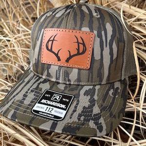 May include: A camouflage trucker hat with a brown leather patch featuring a buck's antlers. The hat has a black label with the text "EST. 1970 RICHARDSON. ORIGINAL 112 TRUCKER ADJUSTABLE".