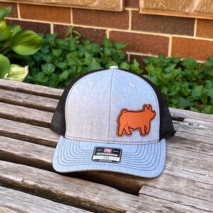 May include: A gray and black trucker hat with a brown leather patch featuring a steer. The hat has a curved brim and a mesh back. The Richardson 112 hat is adjustable.