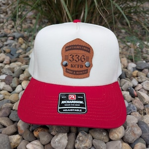 May include: A cream-colored baseball cap with a red brim and a red button on top. A brown leather-like patch shaped like a fire helmet shield is centered on the front, featuring the text BATTALION CHIEF 336 KCFD FIRE & RESCUE.