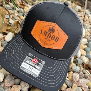 May include: Black and gray trucker hat with a brown leather patch that says "AMDOR FARMS CORNING, IOWA". The hat has a Richardson 112 Trucker label.