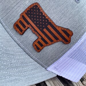 May include: A gray and white trucker hat with a brown leather patch in the shape of a state with an American flag design.
