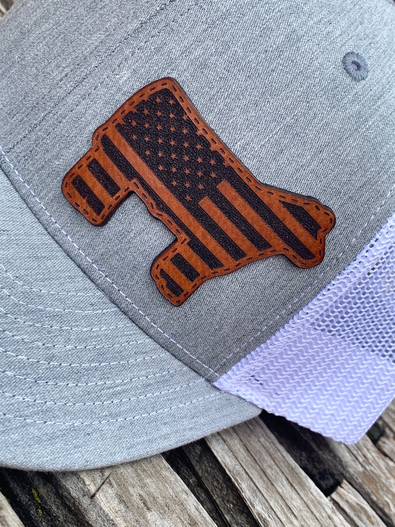May include: A gray and white trucker hat with a brown leather patch in the shape of a state with an American flag design.