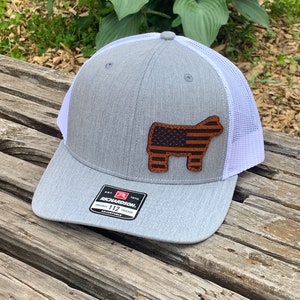 May include: A gray and white trucker hat with a brown leather patch featuring an American flag on a steer silhouette. The hat has a white mesh back and the text "Richardson 112 Original Trucker Adjustable" on the front.