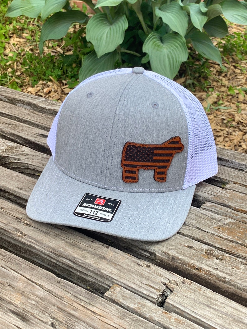 May include: A gray and white trucker hat with a brown leather patch featuring an American flag on a steer silhouette. The hat has a white mesh back and the text "Richardson 112 Original Trucker Adjustable" on the front.