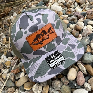 May include: A camouflage trucker hat with a brown leather patch featuring a silhouette of a fish. The hat has a black and white label that reads "Richardson 112 Original Trucker Adjustable".