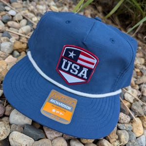 May include: A navy blue baseball cap with a white rope detail and a USA patch. The patch features a star, stripes, and the word "USA" in white. The cap has a flat brim and a "Richardson" label.