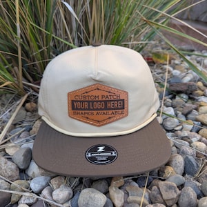 May include: A tan and brown baseball cap with a flat brim. The cap features a leather patch with the text "CUSTOM PATCH YOUR LOGO HERE! SHAPES AVAILABLE." A black and white sticker is on the brim.