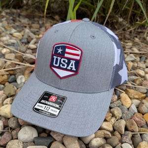 May include: Gray trucker hat with a white and gray camouflage mesh back. The front features a patch with a USA flag design. The hat has a curved brim and an adjustable strap. The tag reads "RICHARDSON ORIGINAL 112 TRUCKER ADJUSTABLE."