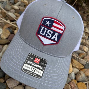 May include: Gray trucker hat with a white mesh back and a USA patch featuring a star and stripes. The hat has a curved brim and an adjustable strap. The Richardson brand tag is visible.