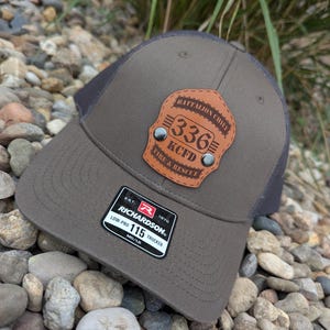May include: A brown and gray trucker hat with a leather patch that reads "BATTALION CHIEF 336 KCFD FIRE & RESCUE." The hat has a Richardson 115 Trucker tag. The hat is sitting on a bed of small rocks.