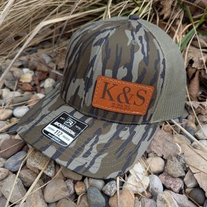 May include: A camouflage trucker hat with a brown leather patch that says "K&S 2.22.25". The hat is made by Richardson 112 and is adjustable.
