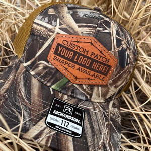 May include: A brown and tan camouflage trucker hat with a brown leather patch that says "CUSTOM PATCH YOUR LOGO HERE! SHAPES AVAILABLE". The hat also has a black and white label that says "EST. 1970 RICHARDSON ORIGINAL 112 TRUCKER ADJUSTABLE".