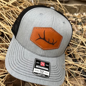 May include: Gray and black trucker hat with a brown leather patch featuring a deer antler design. The hat has a curved brim and a mesh back. The Richardson brand tag is visible.