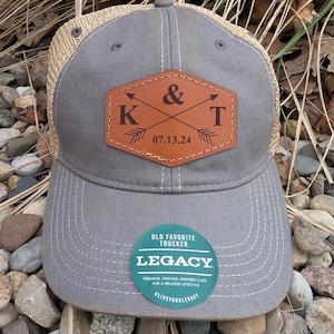 May include: Gray and brown trucker hat with a leather patch that says "K & T 07.13.24" with arrows pointing towards each other. The hat has a mesh back and a Legacy logo on the front.