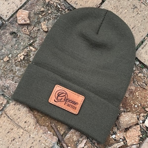 May include: A dark green beanie with a brown leather patch that says "Greene Farms" in a cursive font.