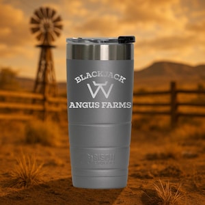 May include: A gray stainless steel tumbler with a black lid. The tumbler has the text "Blackjack W Angus Farms" engraved on the side. The tumbler is sitting on a brown surface.