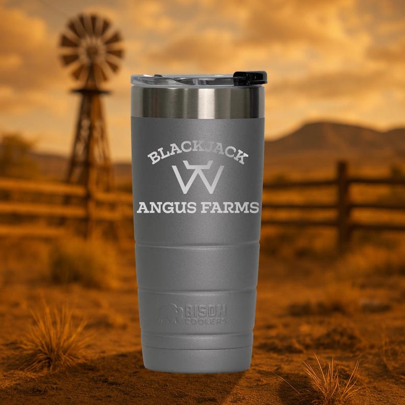 Puede incluir: Un vaso de acero inoxidable gris con tapa negra. El vaso tiene el texto "Blackjack W Angus Farms" grabado en el lateral. El vaso est&aacute; sobre una superficie marr&oacute;n.