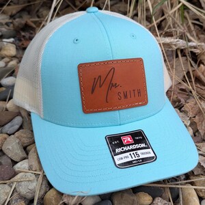 May include: A light blue trucker hat with a white mesh back. The hat has a brown leather patch with the text "Mrs. Smith" in cursive. The hat also has a black and red label with the text "Richardson 115 Low-Pro Trucker MD/LG".