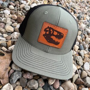 May include: A green and black trucker hat with a brown leather patch featuring a dinosaur skull silhouette.