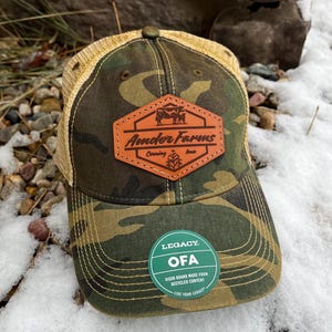 May include: A camouflage trucker hat with a brown leather patch that says "Amder Farms, Corning, Iowa". The hat also has a green circular patch that says "Legacy OFA Visor Board Made From Recycled Content Live Your Legacy".
