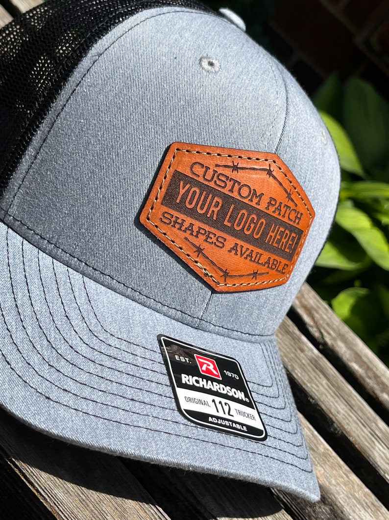 May include: A gray Richardson 112 trucker hat with a black mesh back. It features a brown leather patch with the text "CUSTOM PATCH YOUR LOGO HERE! SHAPES AVAILABLE". The hat is adjustable.