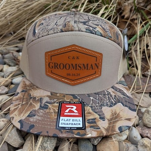 May include: A tan and brown camouflage baseball cap with a brown leather patch that says "C&K GROOMSMAN 08.16.25". The cap has a flat bill and a snap closure. The cap is made by Richardson.