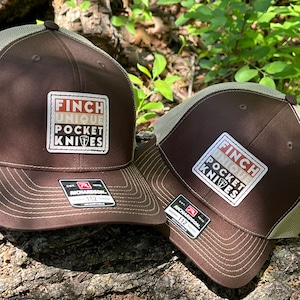 May include: Two brown and beige baseball caps with a mesh back. The caps have a patch on the front that says "Finch Unique Pocket Knives".