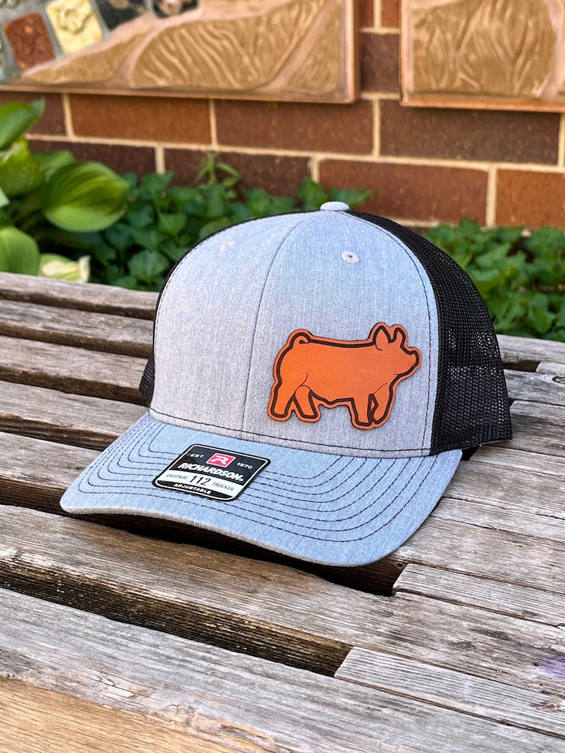 May include: A gray and black trucker hat with a pig patch. The hat has a gray front panel, a black mesh back, and a curved brim. The pig patch is brown and is on the front of the hat. The hat has the Richardson brand label.